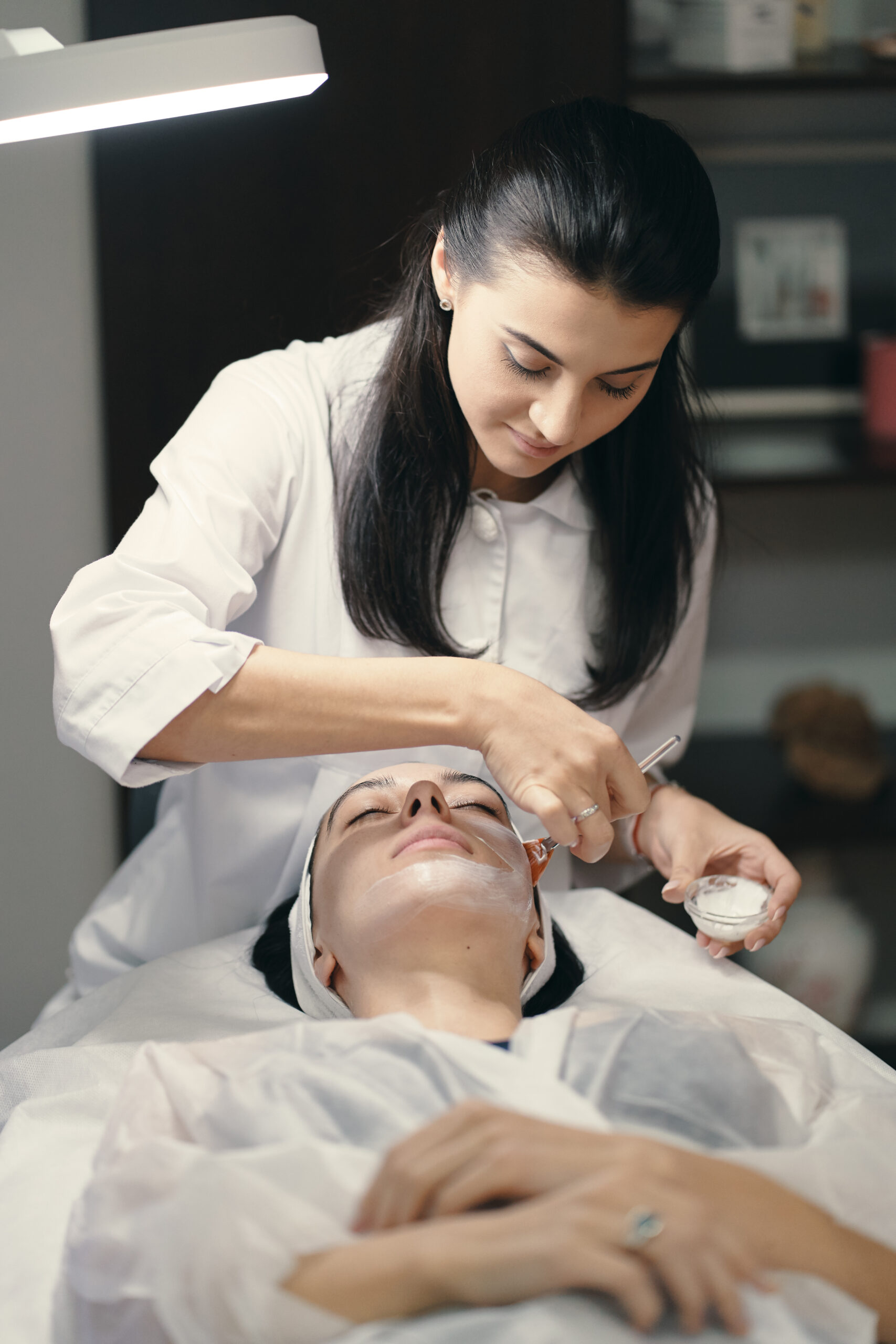 People, beauty, spa, cosmetology and skin care concept - Cosmetologist applying cream on a woman beautiful face with a cosmetologist brush. Lamp light focused on face.
